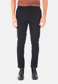Uomo che indossa pantaloni in velluto a coste neri e scarpe di pelle nere, in piedi davanti a uno sfondo chiaro e uniforme.