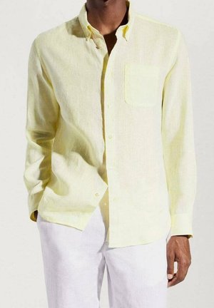 Homme portant une chemise jaune clair à manches longues avec poche poitrine et un pantalon blanc sur un fond clair uni.