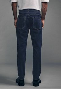 Los jeans de mezclilla azul oscuro presentan un diseño de pierna recta, bolsillos traseros estándar y costuras en contraste. Se llevan con zapatos negros.