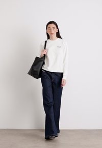 En person går selvsikkert i en hvit hettegenser og mørke jeans, bærende på en svart totebag mot en lys vegg.
