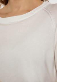 Biała dzianinowa sweter z szerokim dekoltem i prążkowanymi wykończeniami. Materiał wydaje się miękki, o gładkiej teksturze i minimalistycznym wzornictwie.