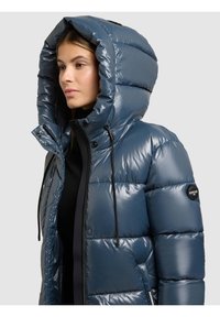 Blå vadderad jacka med glänsande finish, stor huva och dragsko; har en logotyp på ärmen och ribbade muddar.