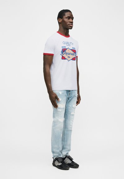 Νεαρός άνδρας στέκεται, φο ρώντας λευκό T-shirt με κόκκινες λεπτομέρειες και γραφικό σχέδιο, ανοιχτόχρωμα σκισμένα τζιν και μαύρα αθλητικά παπούτσια με λευκές ρίγες.