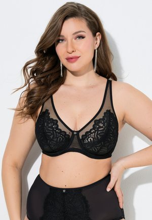 Kvinde med langt, bølget hår, iført sort blonde-bh og matchende højtaljede trusser, poserer med hånden på hoften mod en hvid baggrund.