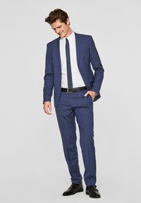 Marineblauer Anzug mit schmaler Passform. Maßgeschneiderte Jacke, passende Hose, weißes Hemd und strukturierte dunkle Krawatte. Schwarze Lederstiefel.