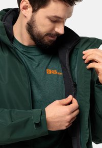 Groene buitenjas met een zachte zwarte voering, ritssluiting en verstelbare manchetten, gecombineerd met een donkergroen T-shirt met een oranje logo.