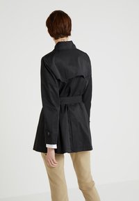 Svart trenchcoat med bälte i midjan, detaljer vid axlarna och knäppte manschetter. Slätt tyg och skräddarsydd passform. Bärs över beiga byxor.