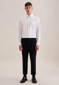 Camicia bianca con bottoni e taschino frontale; abbinata a pantaloni neri su misura e scarpe nere; design e vestibilità minimalisti.