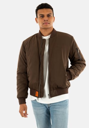 Giacca bomber marrone con chiusura a zip, polsini a coste e un'etichetta arancione. Presenta tasche laterali e un tessuto testurizzato. Indossata sopra una camicia bianca.
