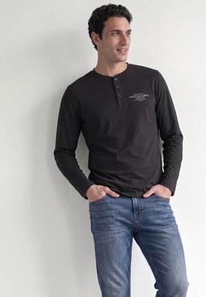 Homme portant un t-shirt henley noir à manches longues avec texte et jean bleu, debout les mains dans les poches, regardant sur le côté, contre un mur blanc.