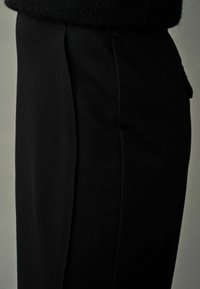 Pantalons noirs ajustés avec un design à jambe droite, présentant une texture lisse et des plis latéraux. La vue en gros plan met en valeur le détail du tissu et la coupe sur mesure.