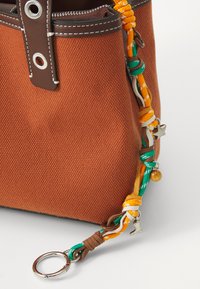 Sac en toile marron avec une sangle tressée colorée ornée de breloques et d'un anneau clé en métal argenté. Couture décorative sur les bords.