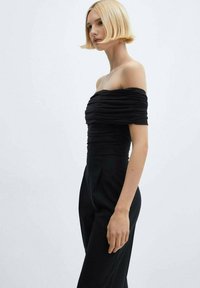 Žena s krátkymi blond vlasmi má na sebe čierny off-shoulder riasený top a čierne nohavice s vysokým pásom na pozadí v svetlej farbe.