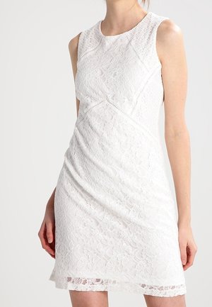 Donna che indossa un vestito bianco senza maniche in pizzo con motivi floreali, in piedi davanti a uno sfondo semplice.