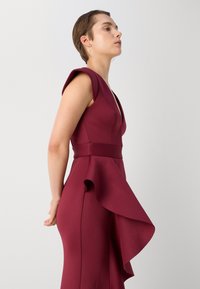 Burgundi spalvos suknelė su prigludusia krūtine, giliu V iškirpimu, trumpomis rankovėmis ir tekančia raukšle palei apačią. Lygus audinys, elegantiška siluetas.