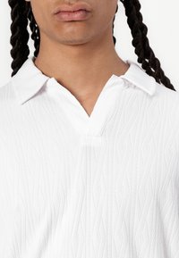 Vit stickad polo t-shirt med ett strukturerat diamantmönster, som har en krage och en delvis knappslå för stängning.