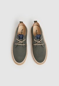 Zapatillas de lona olive con suelas de goma beige, detalles en marrón claro y cordones marrones. Presentan una punta redonda y una plantilla acolchada.