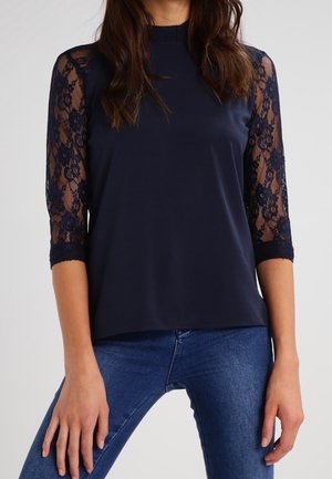 Kvinde iført en marineblå top med florale blonde trekvartærmer og blå jeans mod en ensfarvet baggrund.