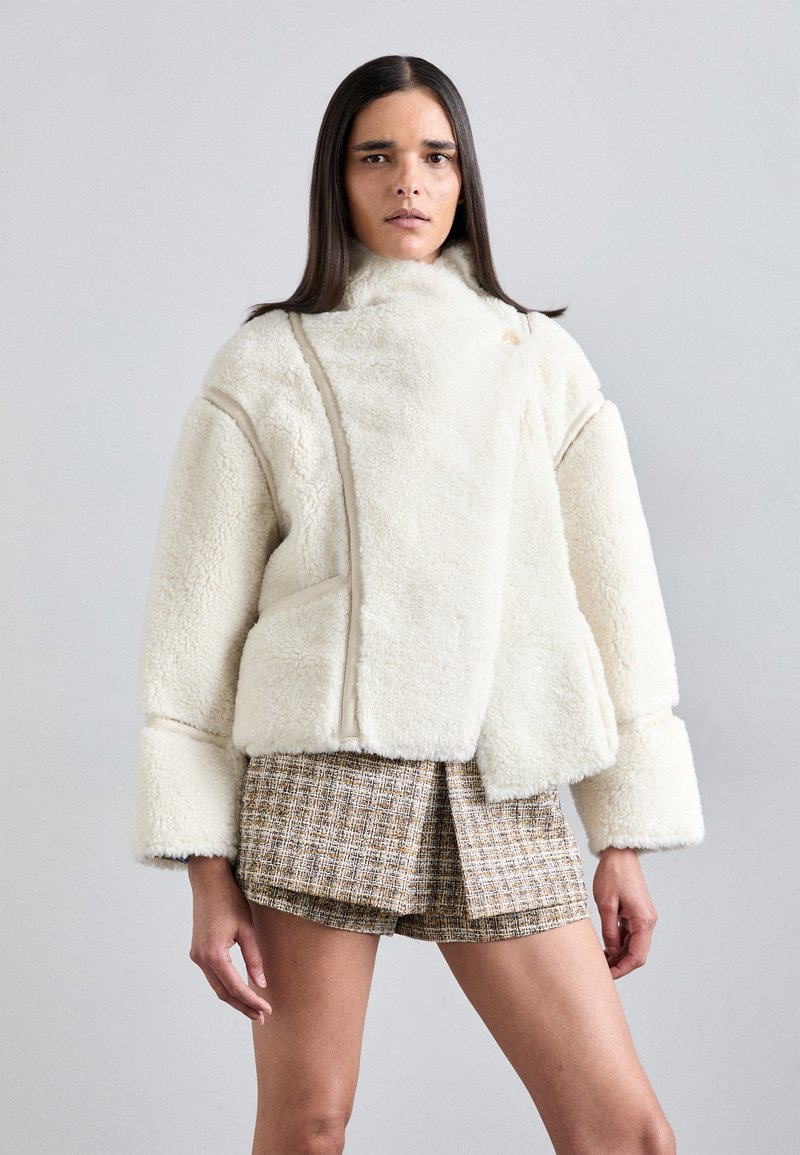 Femeie îmbrăcată cu o jachetă albă pufoasă cu mâneci exagerate și pantaloni scurți din tweed cu model, stând în fața unui fundal gri deschis simplu.