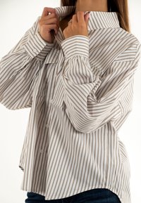 Camisa de botón de manga larga en blanco con finas rayas verticales marrones, que presenta un cuello puntiagudo y mangas fruncidas para añadir volumen.