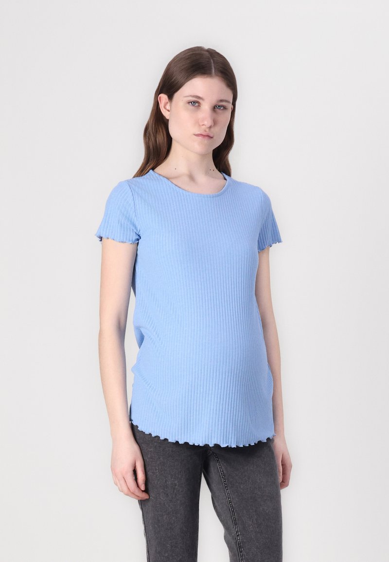 Tricou de maternitate bleu deschis, cu dungi, cu mâneci scurte și volanase și un tiv ondulat, purtat peste blugi strâmți de culoare gri închis.