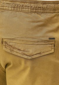 Pantalon en tissu beige avec une poche arrière en biais ornée de surpiqûres contrastantes et une petite étiquette logo en métal près de la couture de la poche. Texture lisse.