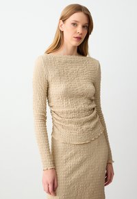 Maglione beige a maniche lunghe con scollatura a barca e dettagli arricciati, abbinato a una gonna coordinata realizzata con un tessuto e una texture simili.