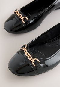 Black patent leather flats with square toes and gold chain detail across the front on a neutral background.