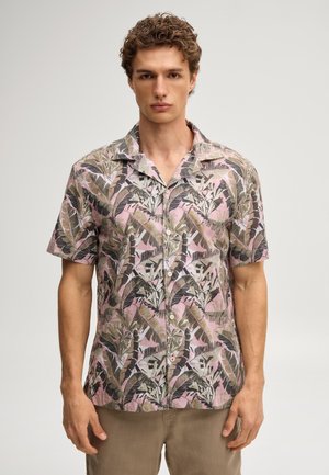 Mann trägt ein kurzärmeliges Hemd mit Knöpfen und einem pink-grünen-schwarzen tropischen Blattmuster, steht vor einem einfarbigen Hintergrund.
