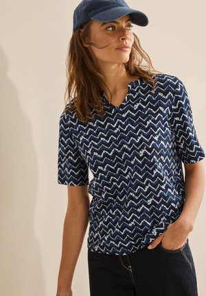 Camicetta a maniche corte in blu navy con un motivo a zigzag bianco, caratterizzata da un collo a V e vestibilità comoda, abbinata a jeans scuri e un cappellino blu.