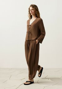 Femme aux cheveux ondulés portant un haut marron texturé à manches longues ample et un pantalon assorti, une chemise blanche en dessous, et des sandales noires, debout sur un sol en pierre.