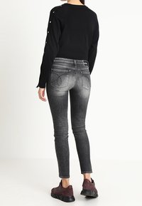 Black sweater with button details on sleeves, paired with fitted gray denim jeans featuring back pockets and a faded wash, worn with burgundy sneakers.