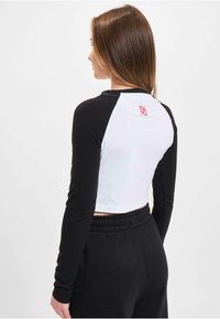 Top cropped cu mâneci lungi, alb-negru, cu guler rotund; prezintă un logo roșu pe spate și mâneci ajustate. Material texturat.