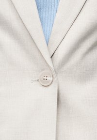 Φωτεινό μπεζ σακάκι με κουμπί πάνω από ένα ανοιχτό μπλε πλεκτό πουλόβερ, που δείχνει την υφή του υφάσματος και τη λεπτομέρεια του κουμπιού στο μπροστινό μέρος.