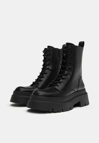 Fekete bőr bokacsizma vastag talppal, fűzős elejével és oldalsó cipzárral. Enyhén lekerekített orr és struktúrált talpdesign.