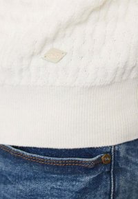 Maglione bianco lavorato a maglia con un design testurizzato e un'etichetta tessuta. Abbinato a jeans in denim con un accento di cuciture arancioni.