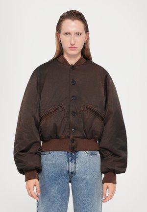Veste bombardier marron avec de grandes manches bouffantes, poignets et ourlet côtelés, dotée de boutons sur le devant et de deux poches latérales. Portée avec un jean bleu clair.