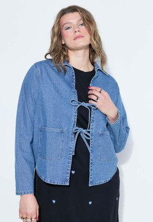Junge Frau mit welligem blondem Haar, die eine blaue Jeansjacke mit vorne gebundenen Schleifen über einem schwarzen Kleid mit kleinen blauen Herzen trägt.