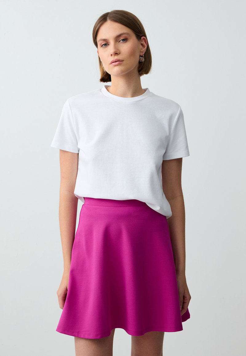 T-shirt bianco in cotone a maniche corte, abbinato a una gonna rosa svasata e lunga fino al ginocchio, realizzata in un tessuto liscio e elastico. Senza motivi.