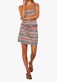 Robe grise à motifs avec des rayures orange et blanches, bretelles réglables et taille cintrée. Portée avec des tongs noires.