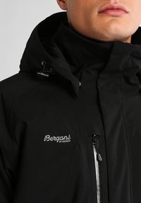 Czarna wodoodporna kurtka z wysokim kołnierzem, regulowanym kapturem, zapięciem na zamek z przodu oraz haftowanym logo na piersi.