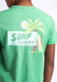 Zelené bavlněné tričko s bílým obdélníkovým grafickým vzorem, na kterém je nápis "SURF RIDERS", ilustrace palmy, surfového prkna a slunce v žluté a růžové barvě.
