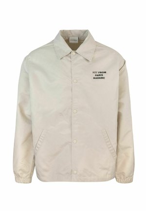 Giacca leggera beige con colletto a punta, bottoni a pressione, tasche laterali e testo nero "NON DA PARIGI MADAME" sul petto.