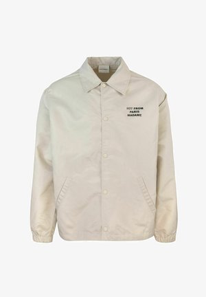 Giacca leggera beige con colletto a punta, bottoni a pressione, tasche laterali e testo nero "NON DA PARIGI MADAME" sul petto.