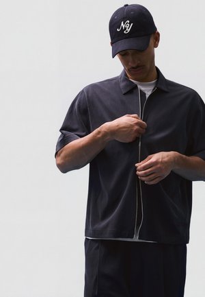 Mand iført en mørk lynlås-skjorte og matchende bukser, som lynlåser jakken, med en sort kasket med hvid "Ny"-skrift.