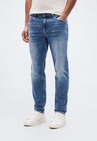 Τζ ιν παντελόνι ανοιχτό μπλε με ίσια γραμμή, με ελαφρύ ξεφλούδισμα, πέντε τσέπες και κούμπωμα με κουμπί, συνδυασμένο με λευκά αθλητικά παπούτσια.