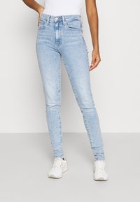 Vaaleansiniset farkut, valmistettu denimistä, joissa on korkea vyötärö ja perinteinen viiden taskun mallisto, yhdistettynä valkoisiin tennareihin.