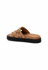 Sandalias de piel de ante marrón con tachuelas en tono dorado y gemas falsas, que cuentan con una suela plana de goma negra y un diseño de punta abierta.