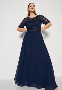 Rochie de seară bleumarin cu un corp din dantelă, mâneci scurte și un brâu cu paiete. Fustă din chiffon fluidă cu o ușoară formă A.