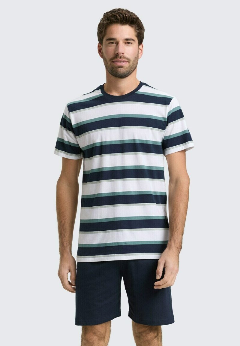 Man draagt een korte mouw T-shirt met ronde hals met horizontale strepen in marineblauw, wit en teal en effen donkere korte broek tegen een effen achtergrond.